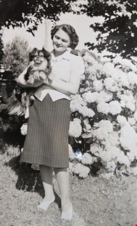 Hazel Simnett standing in a garden, with a small dog in her arms [194-]. Item no. 549-063. thumbnail